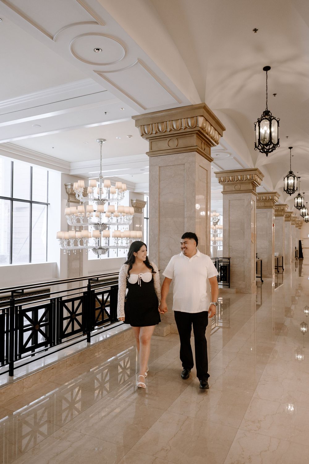 Two people walk together in a luxurious corridor with ornate architectural details and lighting fixtures.