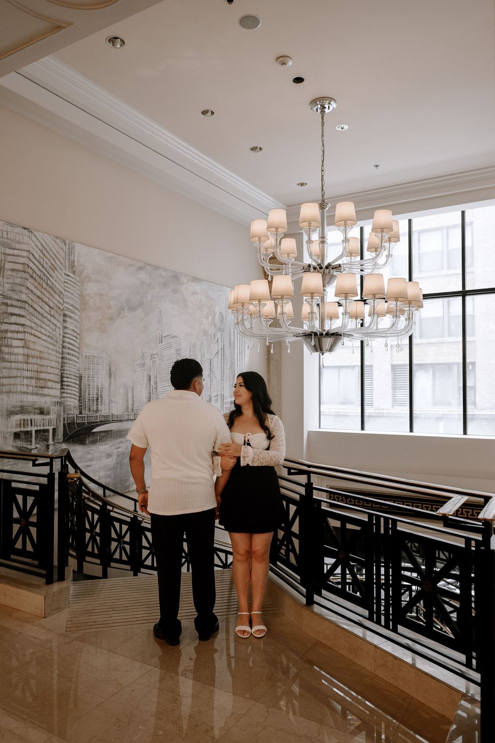 Couple admiring elegant interior with chandelier and black railings while standing together in formal attire.