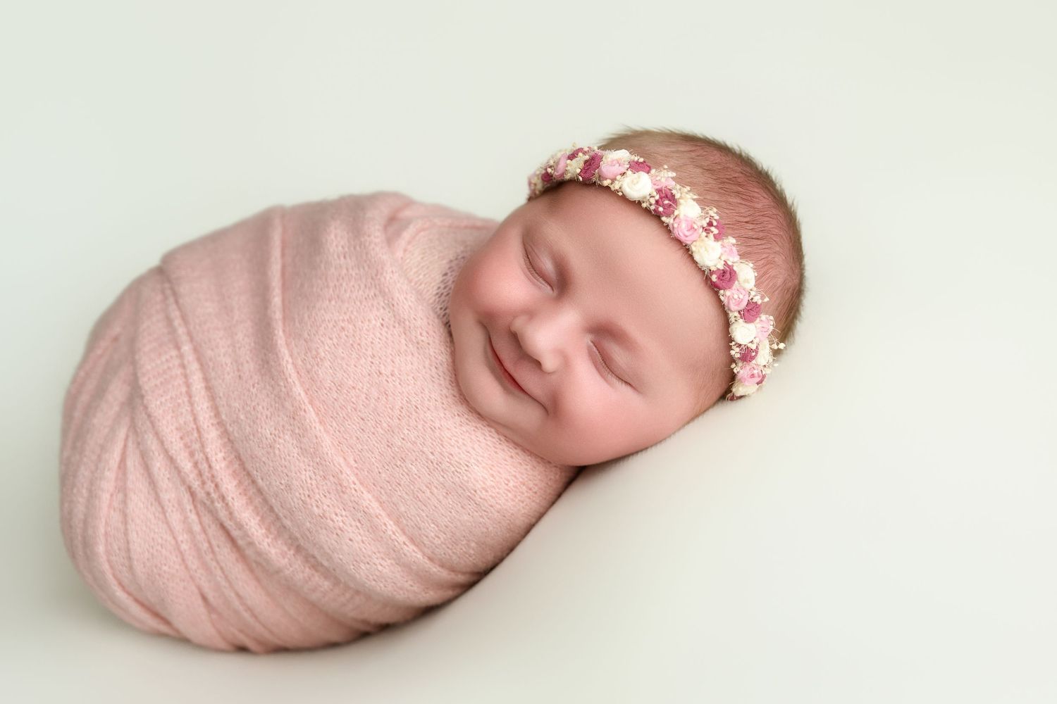 Sleeping newborn baby wrapped in soft pink swaddle wearing delicate floral headband smiling during professional photoshoot.