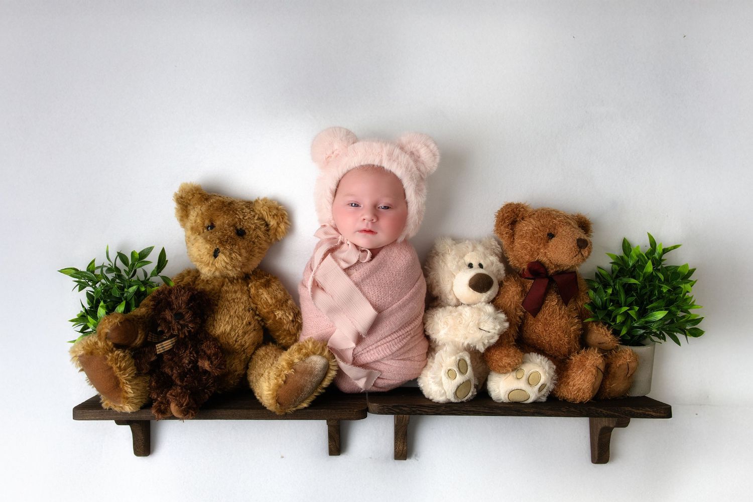 A small baby wearing a pink bear hat sits between four plush teddy bears on a wooden shelf with greenery decorations.