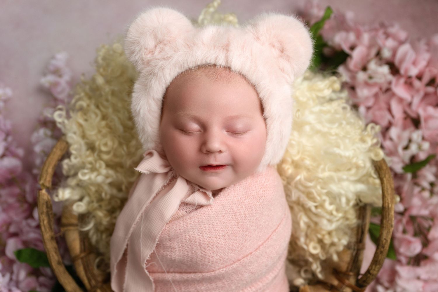 A newborn baby wearing white sleeps peacefully surrounded by soft pink blossoms and delicate cream flowers.