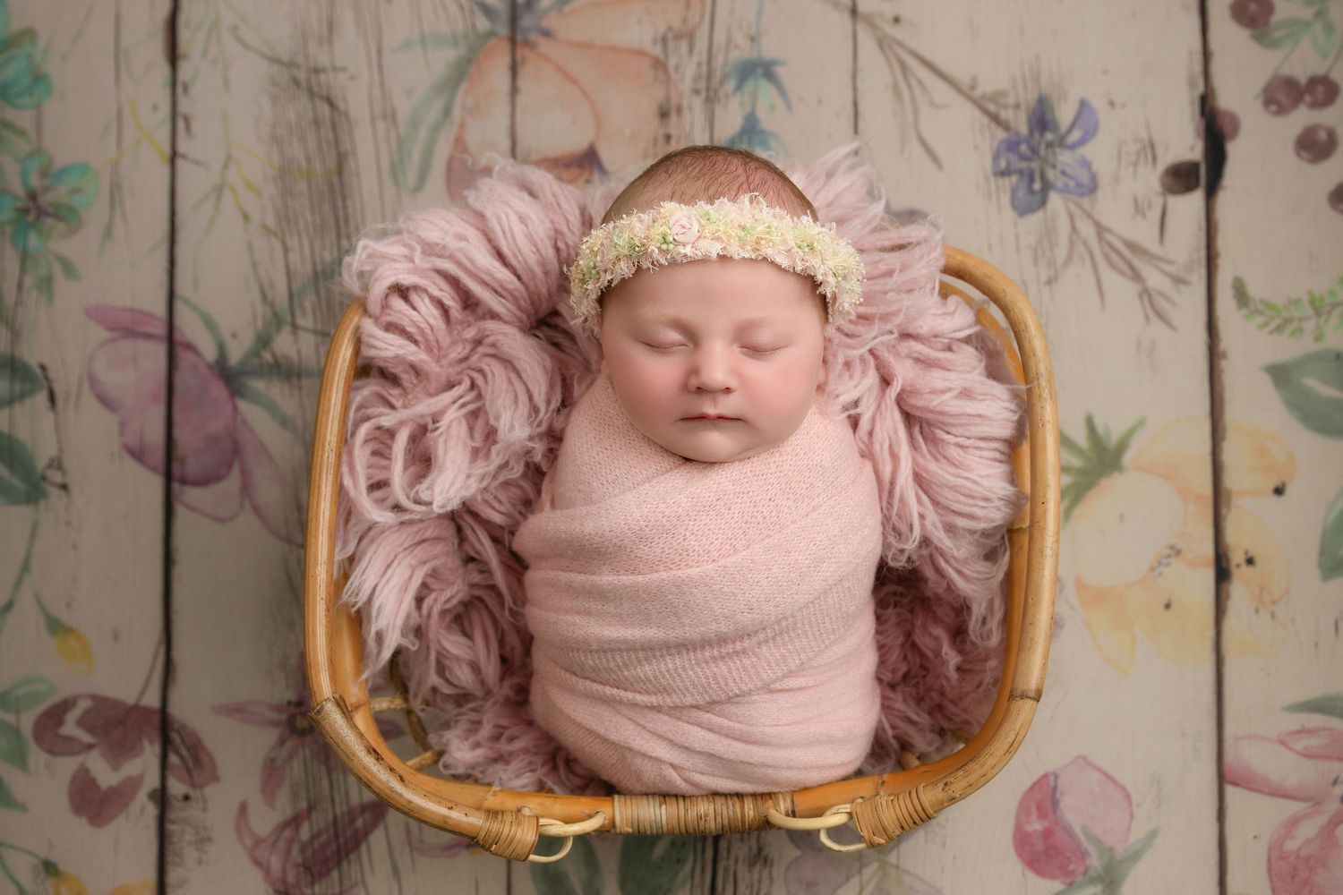 Sleeping newborn wrapped in pink swaddle nestled in basket with floral backdrop.