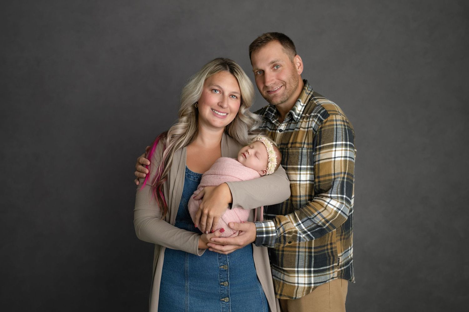 Family portrait of three against dark gray background with parents wearing casual clothing holding infant.