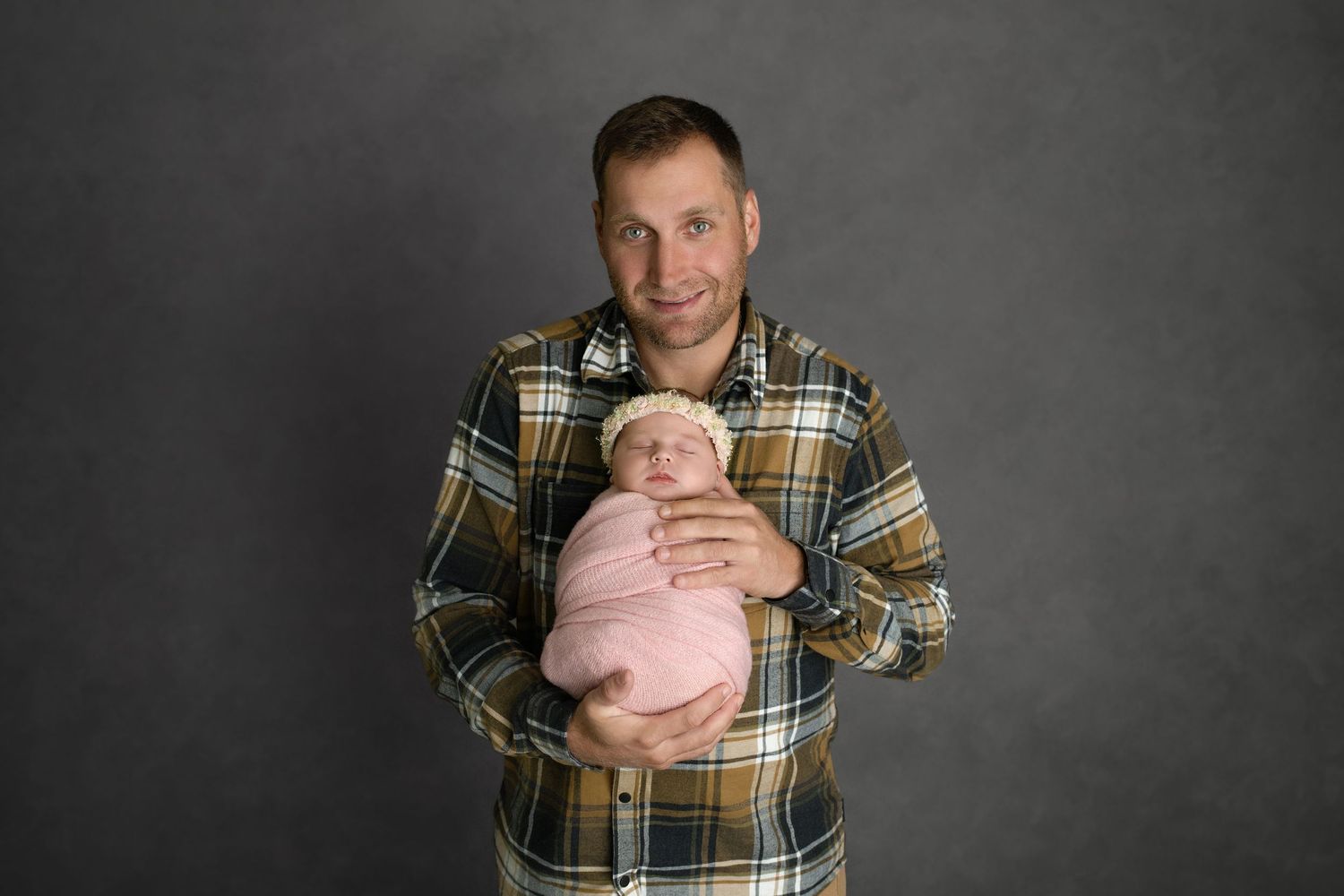 Adult wearing plaid flannel shirt holds sleeping newborn baby swaddled in pink against grey studio backdrop.