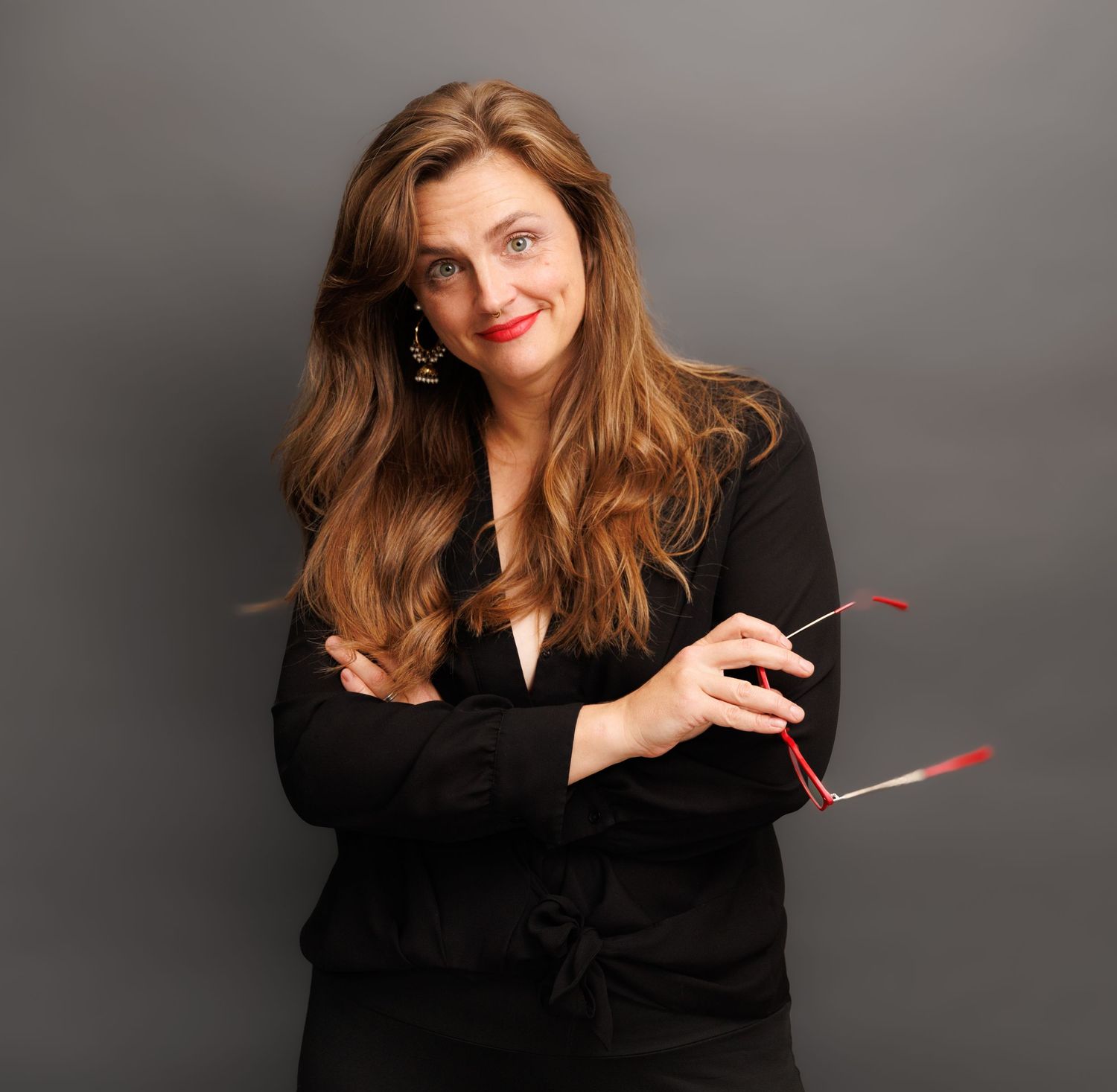 Smiling woman with red hair holding glasses, wearing black blouse against gray background.