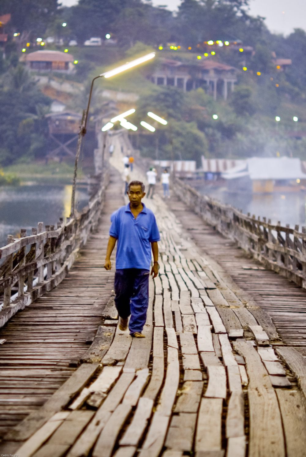 A rickety wooden footbridge spans across water with streetlights visible and houses on a hillside in the background.