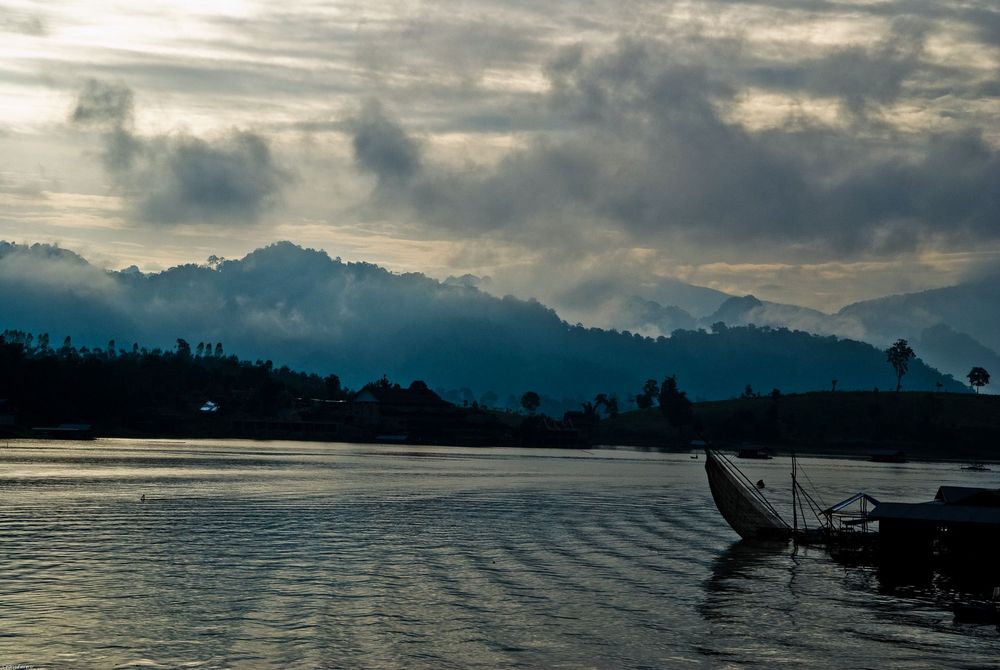 Silhouette of a traditional boat on tranquil lake waters against misty mountain backdrop during golden hour.