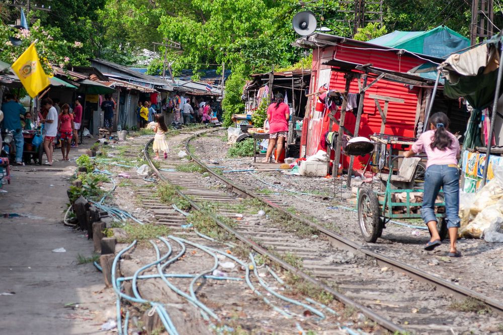 Overgrown train tracks wind through a bustling local market with colorful stalls and vendors along both sides.
