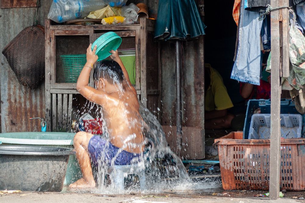 Person pouring water with a bucket during bath or shower outside a rustic building with hanging laundry.