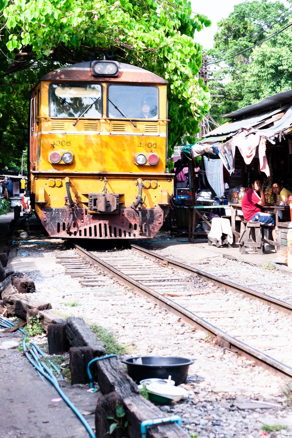 Yellow train engine parked on railway tracks at a rural station with lush green trees in the background.