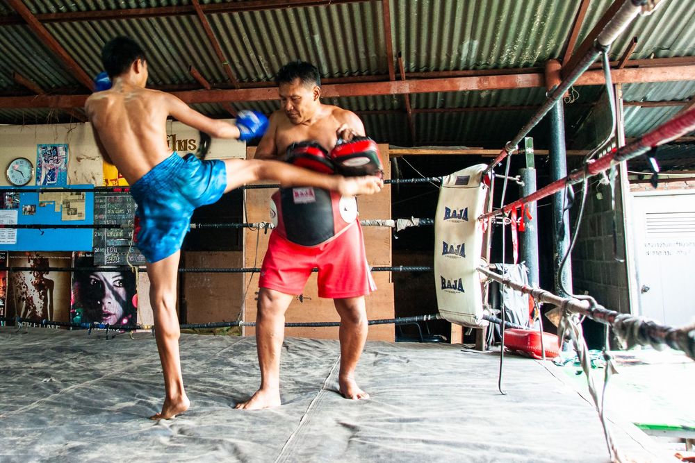 Muay Thai fighters practicing traditional techniques and stretches along a covered outdoor gym training area.