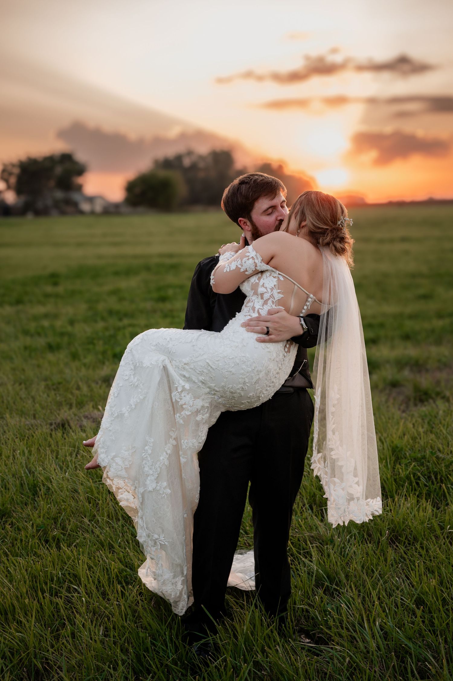 Groom lifting bride in white lace gown and veil, sharing a kiss in a green field at golden sunset.