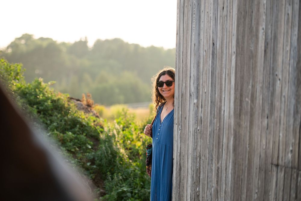Woman in blue dress and sunglasses poses against wooden wall during golden hour sunset.