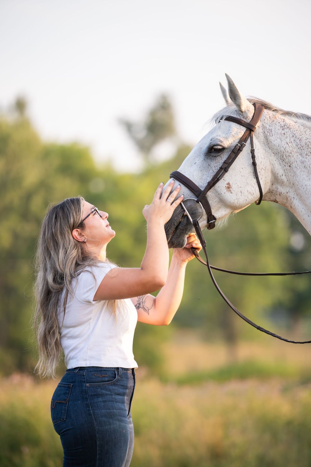 A white horse and someone in a white shirt share a playful moment in a sunny outdoor field.