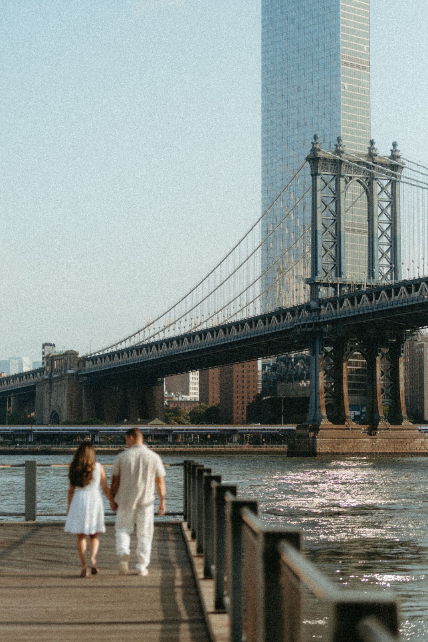 Two people walk along the East River promenade with the Manhattan Bridge in the background on a sunny day.
New York engagement photographer