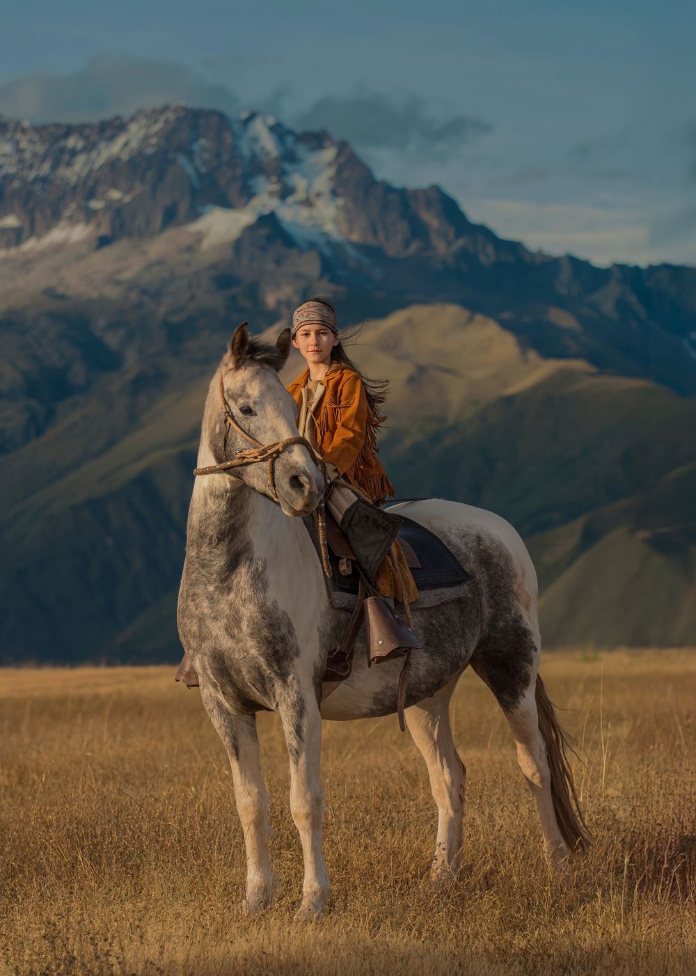 A dappled grey horse and rider traverse a golden grassland with dramatic mountain peaks rising in the background.