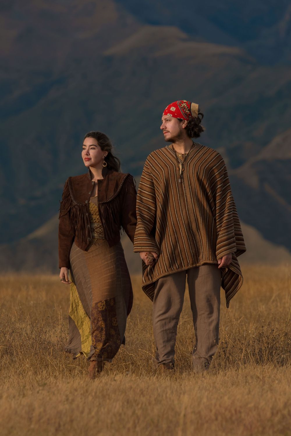 Two figures in traditional ponchos walk together through a golden wheat field against a mountainous backdrop at sunset.