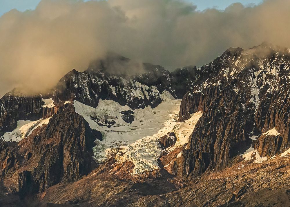 A panoramic view of rugged mountain peaks with snow patches and dramatic clouds during golden hour.