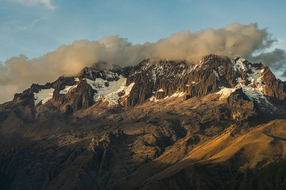 Rugged mountain peaks catch golden sunlight against a blue sky in a panoramic landscape view.