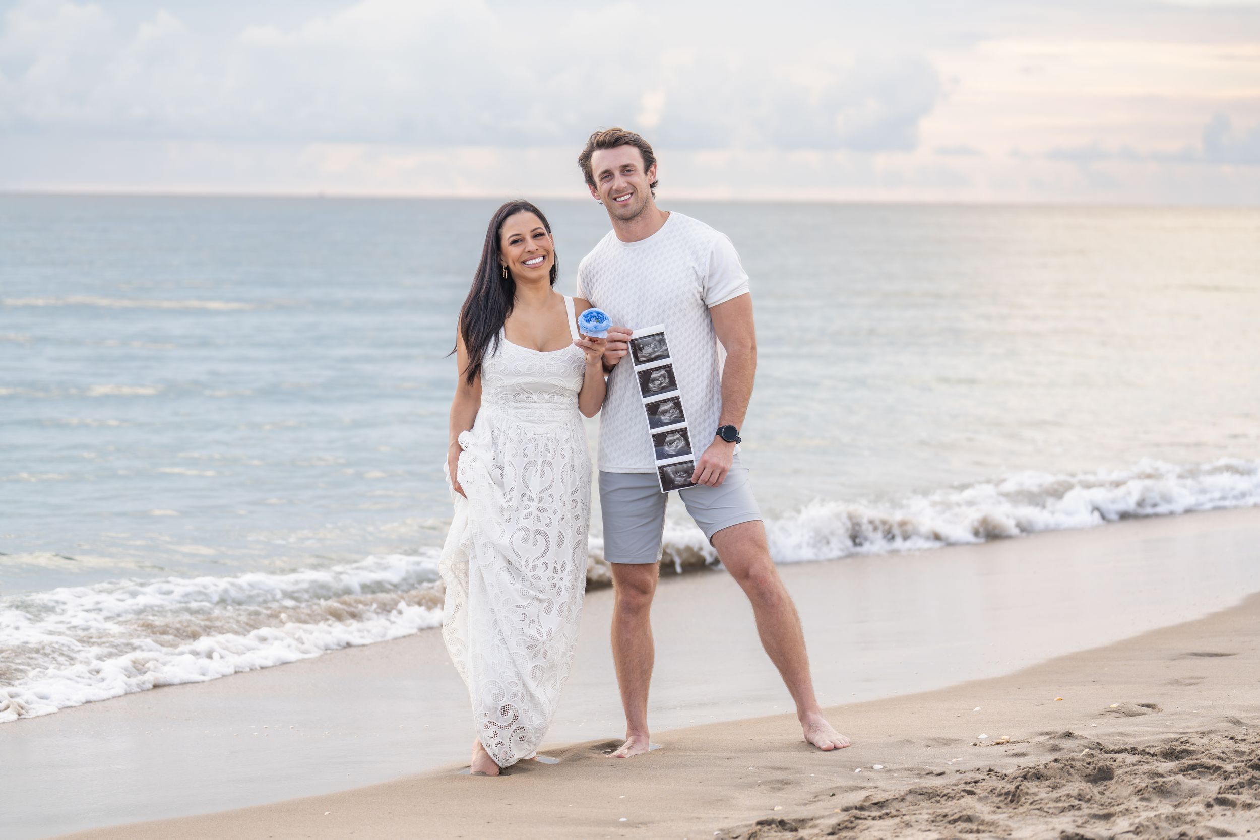 Couple walking together on a scenic beach at sunset holding hands and smiling in casual white outfits.