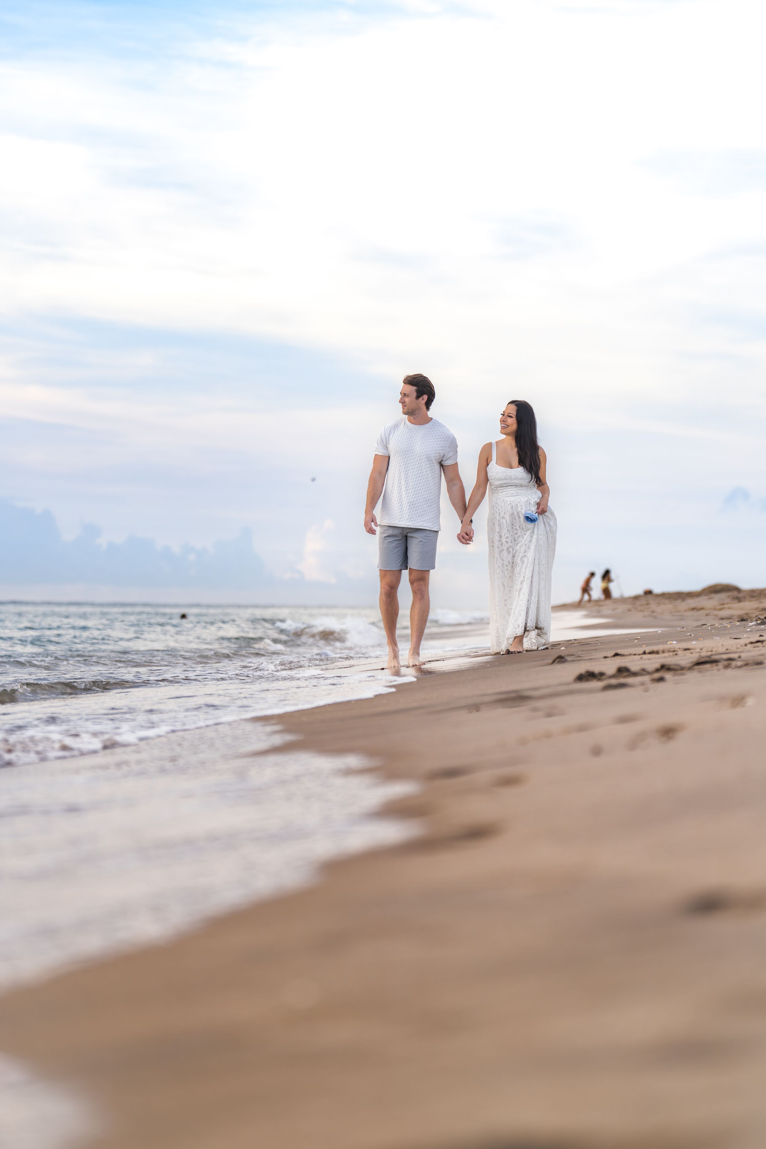 Barefoot couple strolling along sandy beach with ocean waves in the background during golden hour.