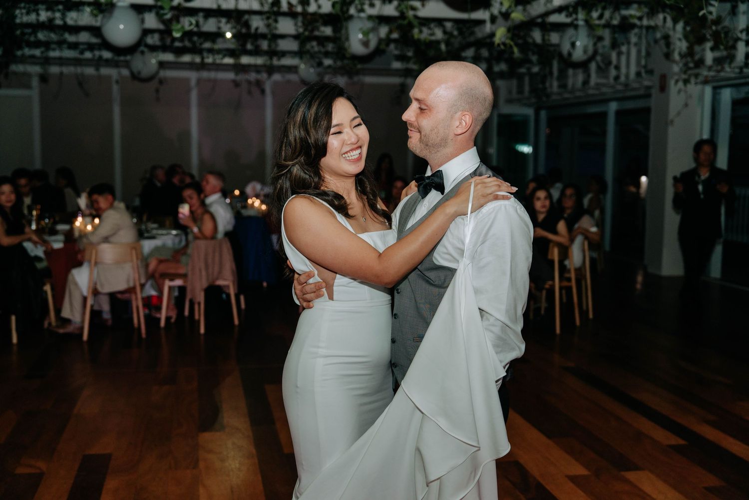 A couple dances together during a wedding reception on a dark wooden dance floor.
