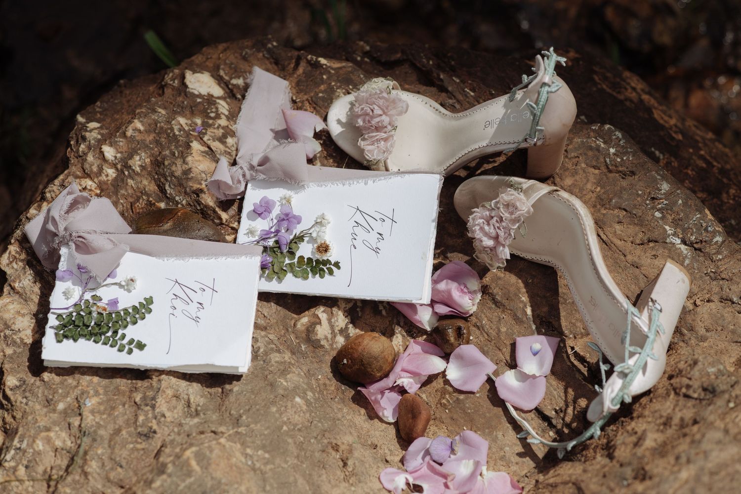 White bridal heels beside botanical-illustrated wedding cards and scattered flower petals on rocky surface.