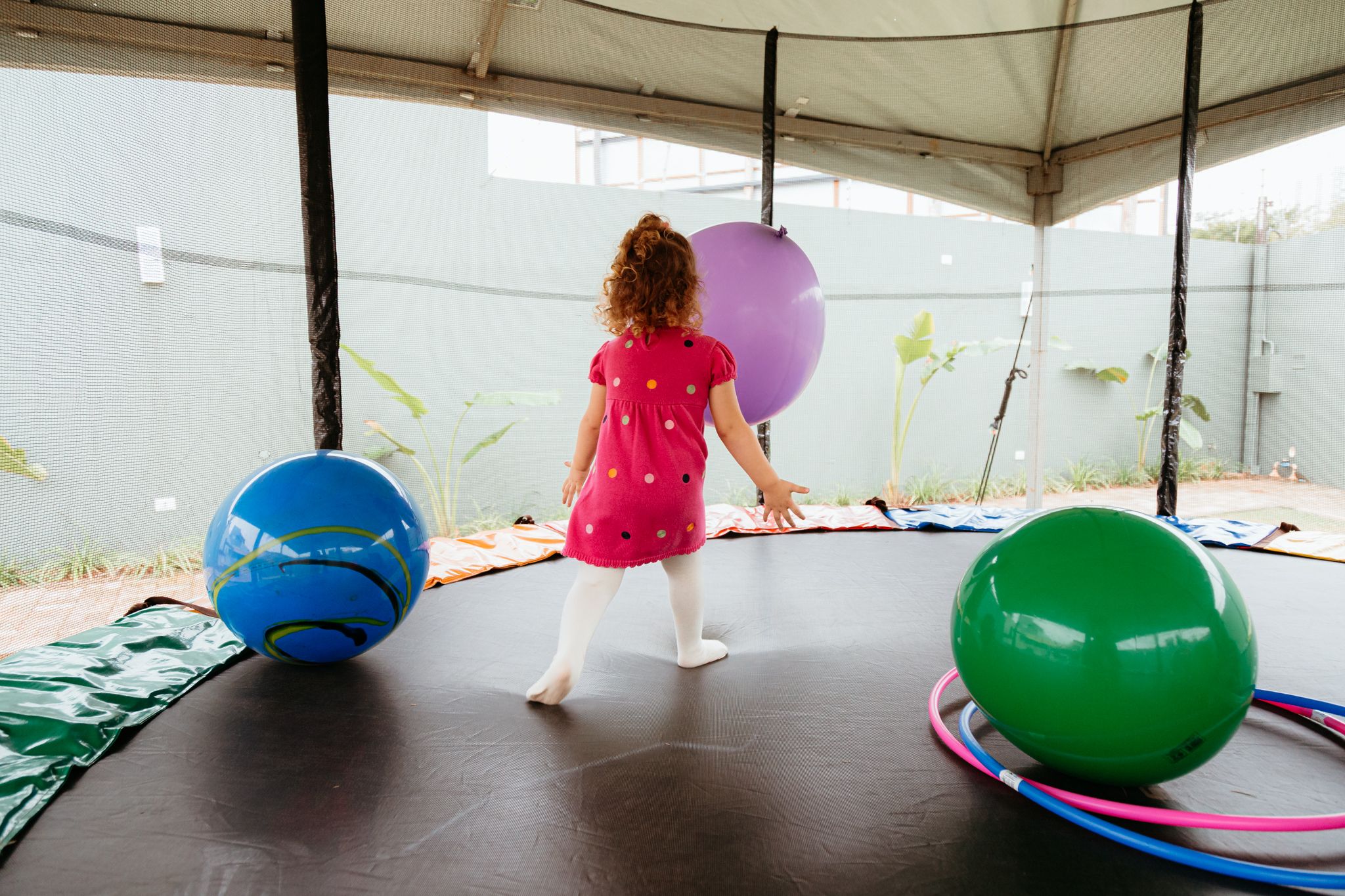 Child in pink dress playing on trampoline with colorful exercise balls inside covered outdoor space.