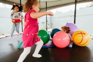 Children playing on a trampoline with colorful balloons during an indoor party or activity session.