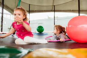 Children in colorful clothing play with large red and orange exercise balls in an indoor activity space.