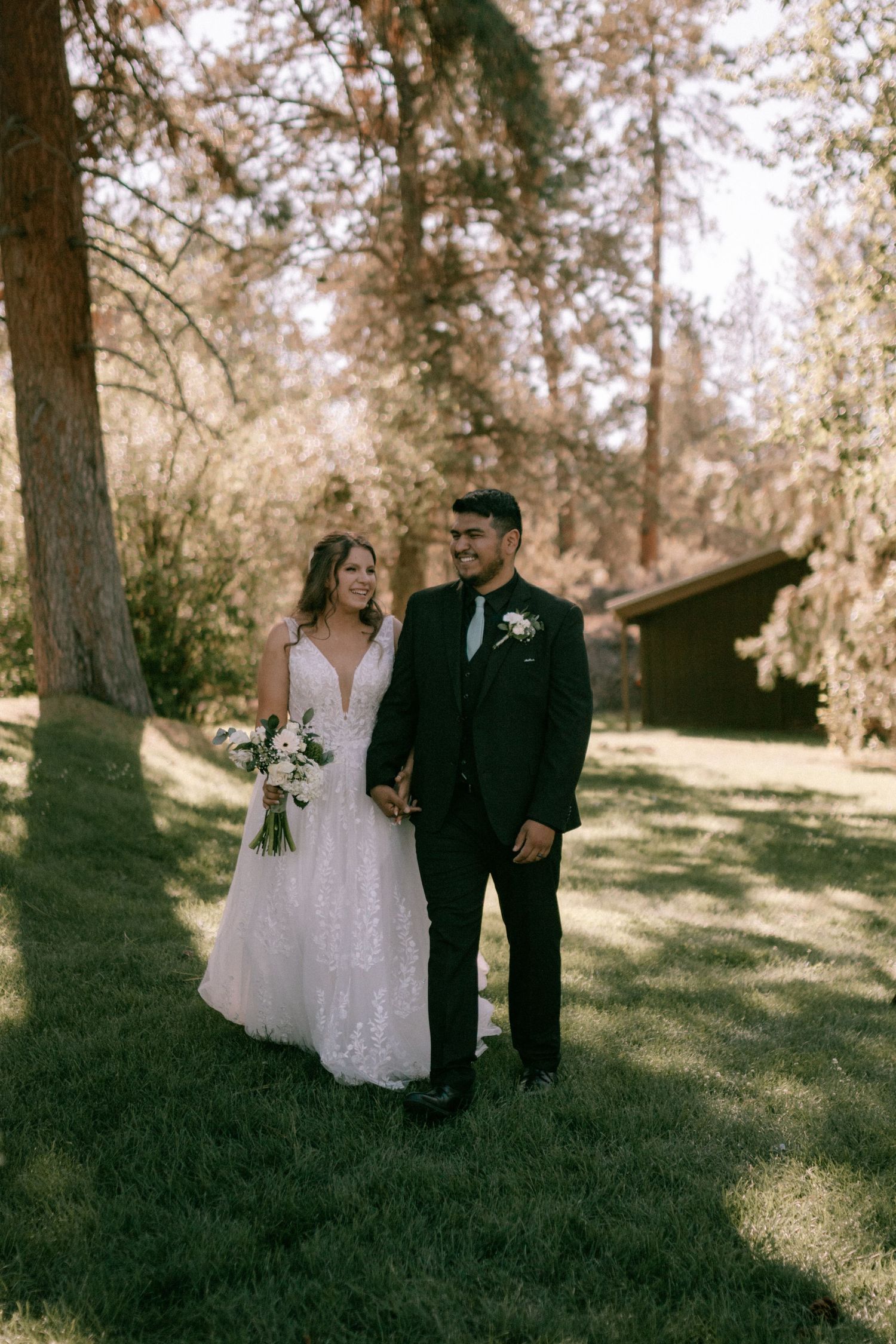 Newlywed couple walking together on a sun-dappled grassy hillside surrounded by trees during an outdoor wedding ceremony.