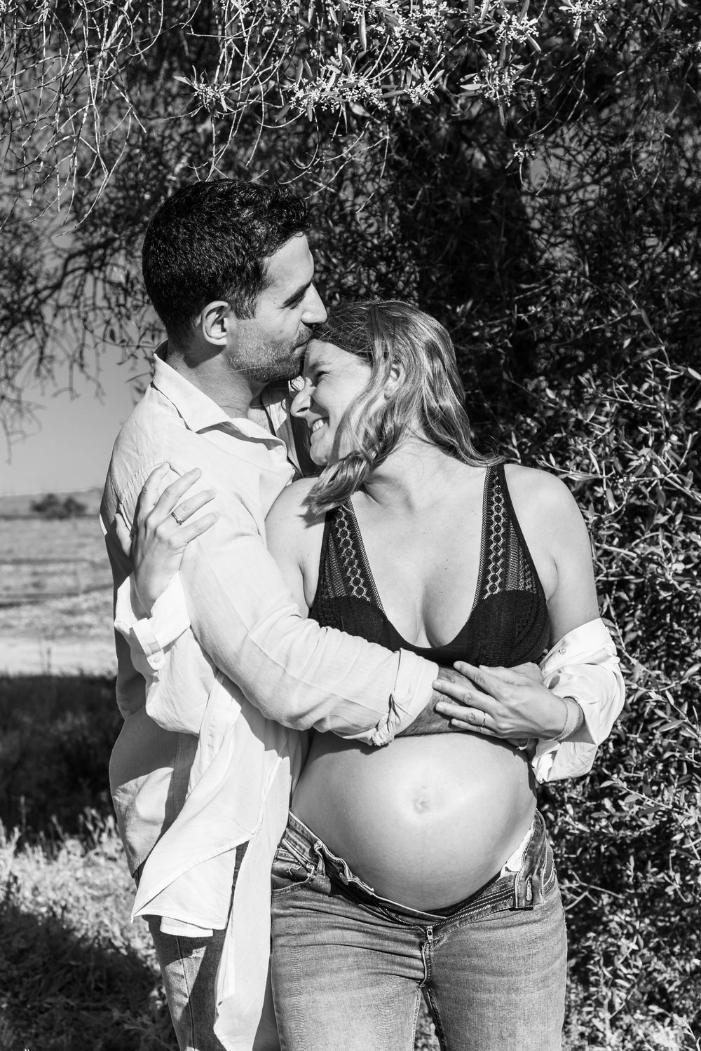Black and white romantic maternity photoshoot showing tender couple moments outdoors near foliage.