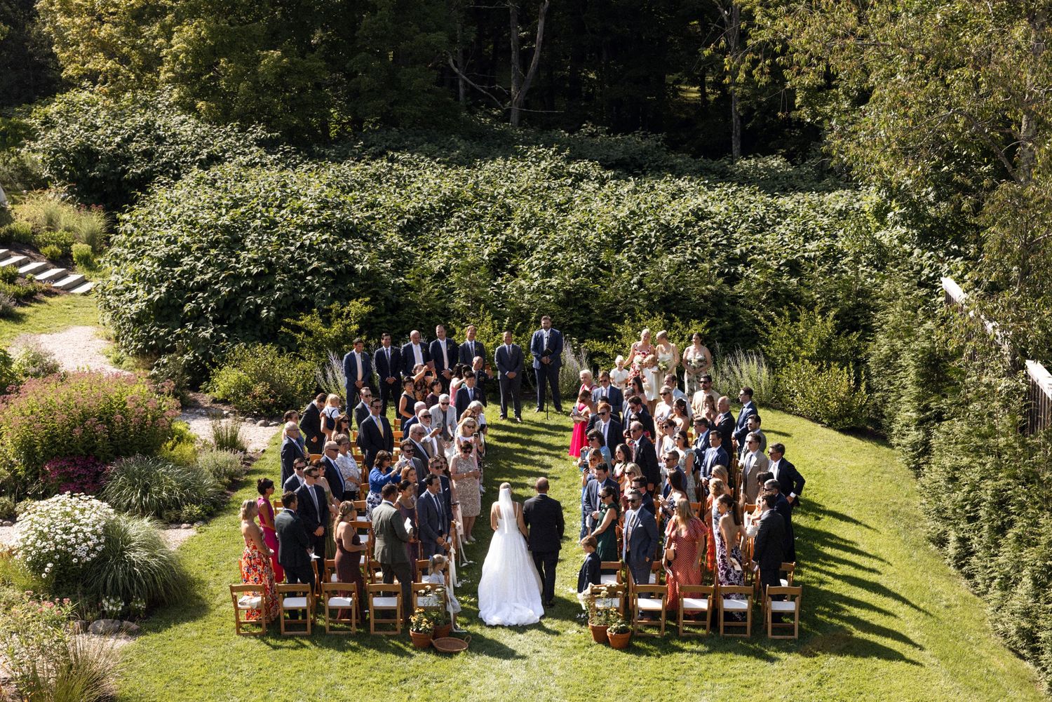 Panoramic view of wedding guests seated for an outdoor ceremony surrounded by lush gardens.