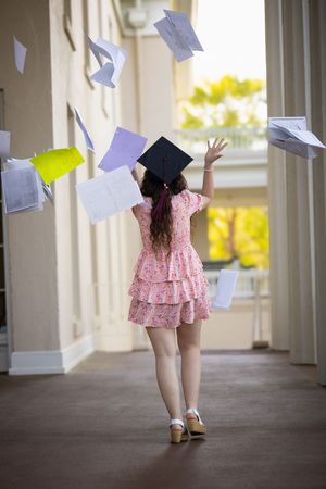Graduate wearing pink dress throws papers in the air while walking down a covered walkway on graduation day.