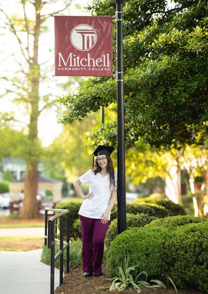 Student in graduation regalia poses with Mitchell College signage outdoors on a sunny spring day.
