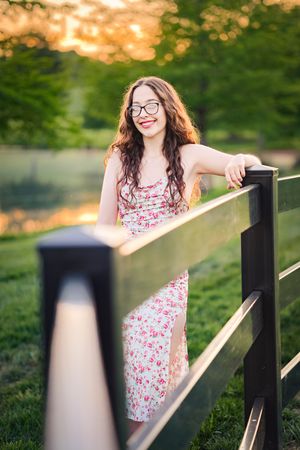 A person in a floral dress poses along a black railing at sunset with green grass in the background.
