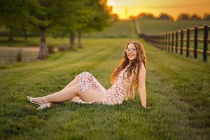 Graduate in pink dress reclines on grassy hill during sunset photoshoot with black academic gown.