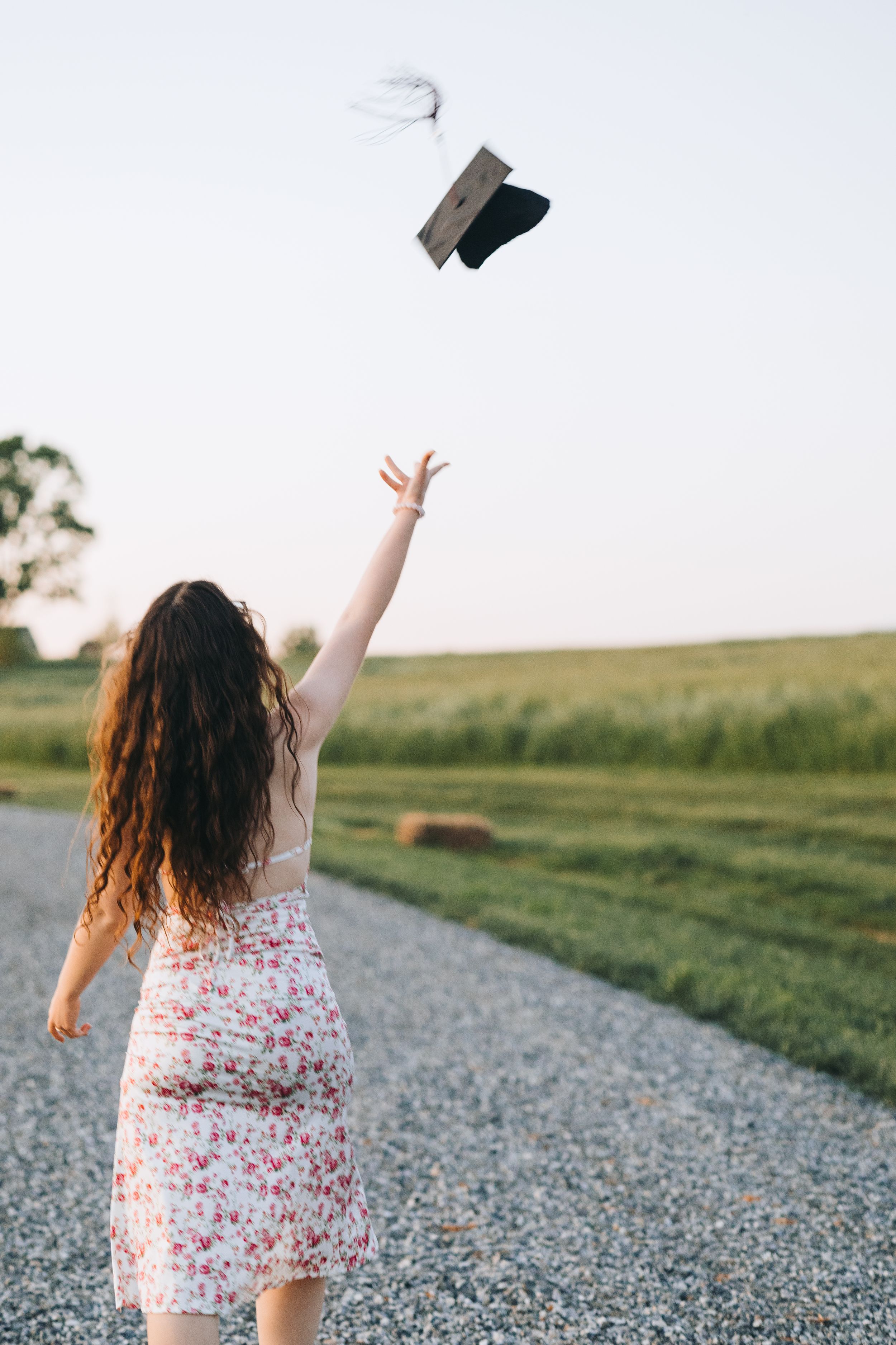Person in a floral dress throws papers into the air while standing on a gravel path surrounded by green fields at sunset.