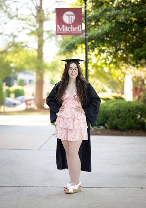 Graduate in black graduation gown and pink outfit poses on campus walkway during golden hour.