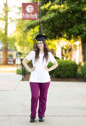 Graduate in white top and purple pants poses on university campus walkway wearing graduation cap.