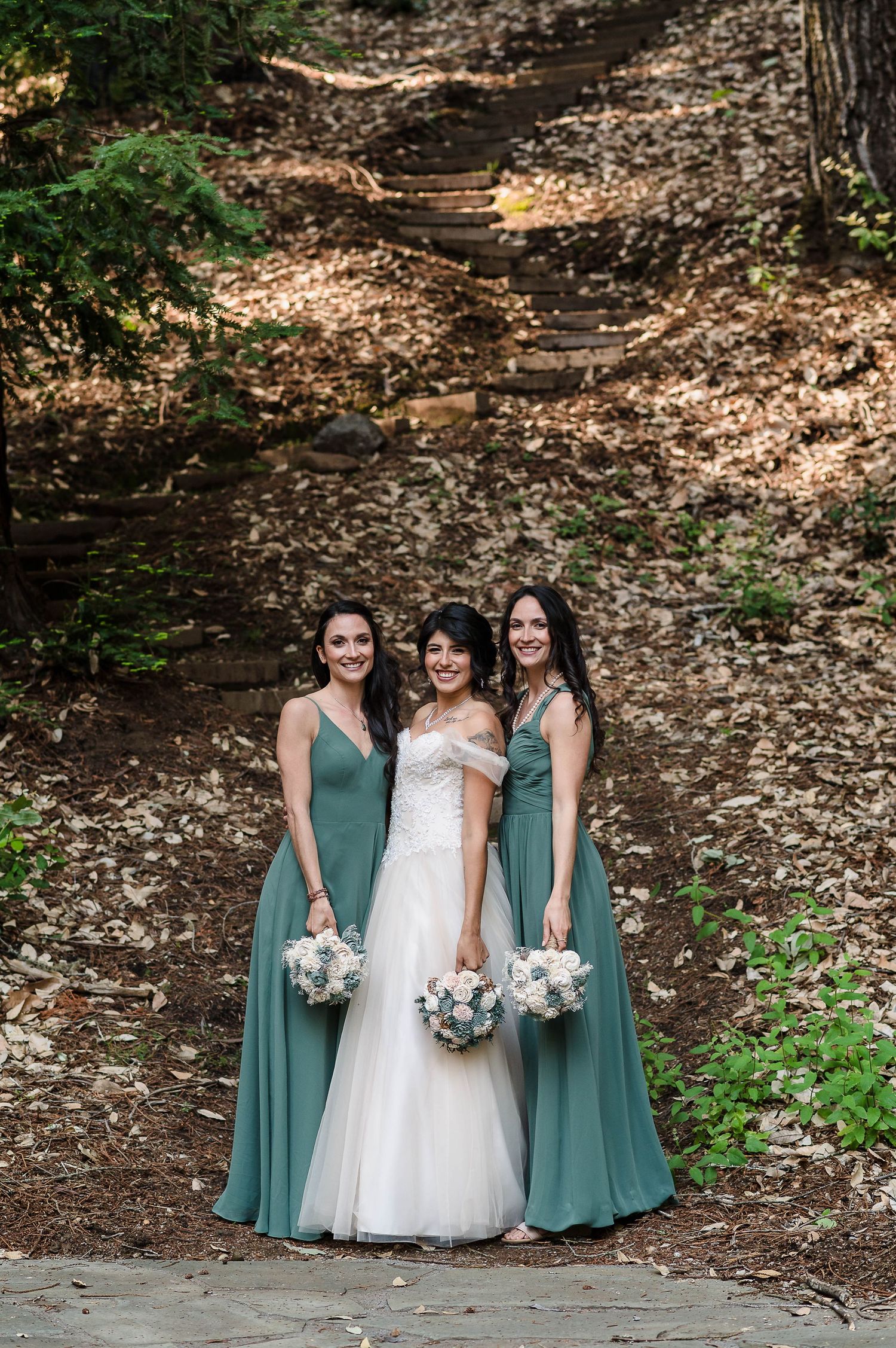Bridesmaids wearing sage green dresses pose with bride in white gown on a wooded path during outdoor wedding photography.