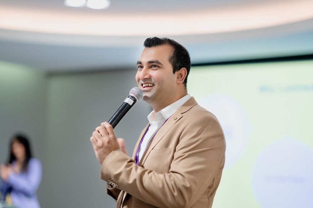 Professional speaker in beige suit jacket presenting at business conference with microphone and screen display.