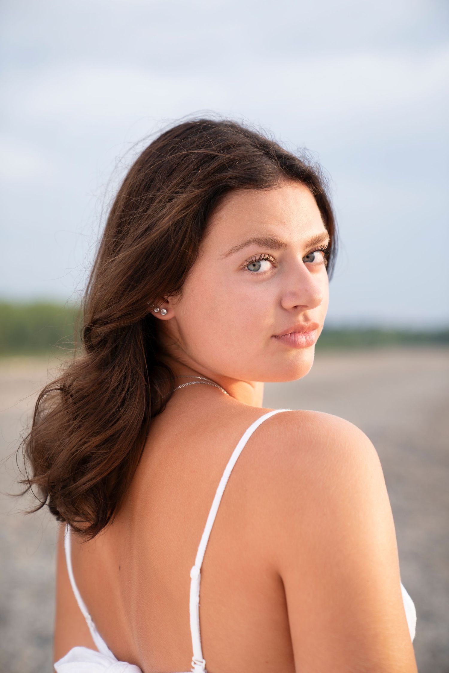 A young woman with dark hair in a white tank top glances over her shoulder against a soft blurred background outdoors.