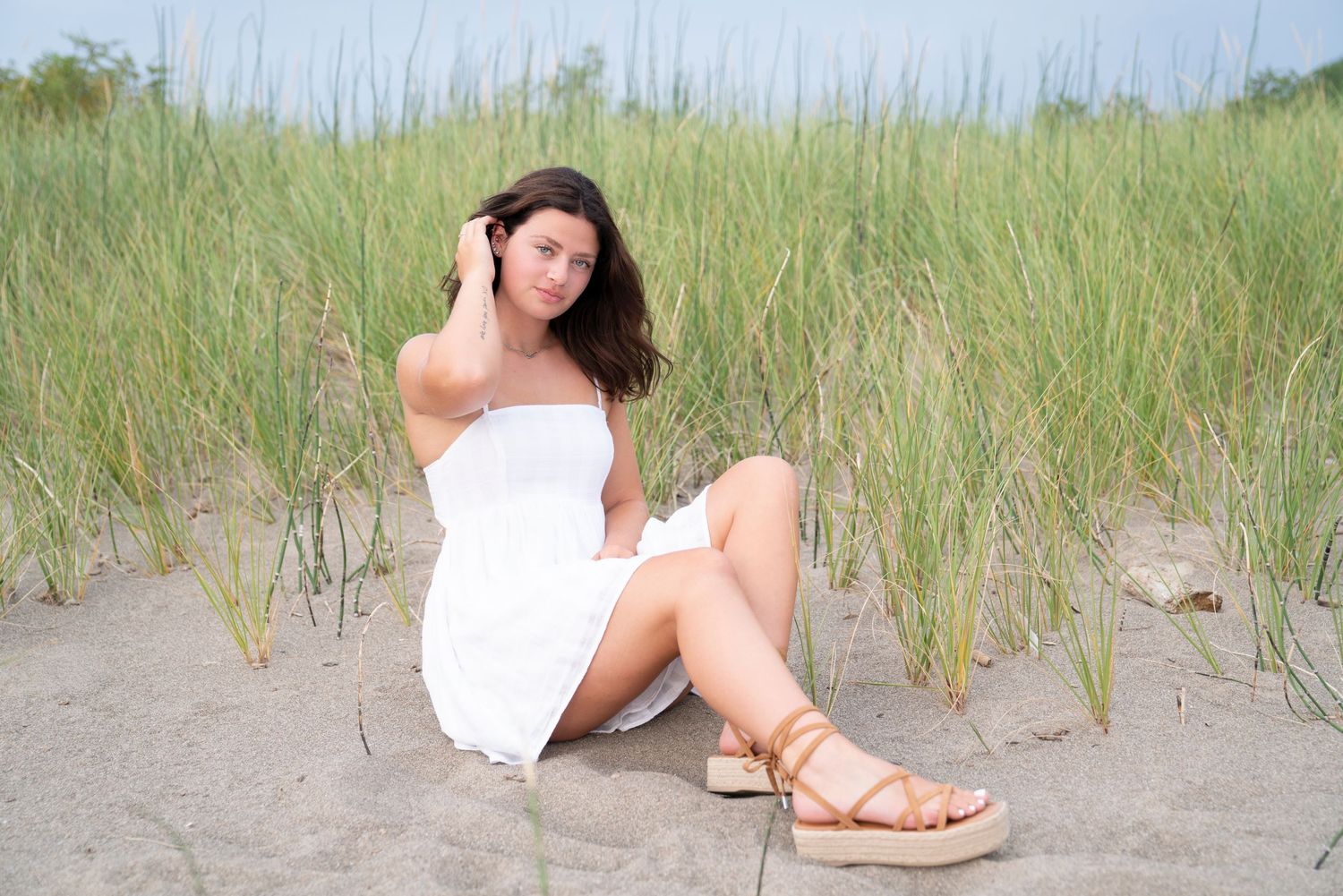 Someone in a white sundress relaxes among tall beach grass and sand dunes on a summer day.