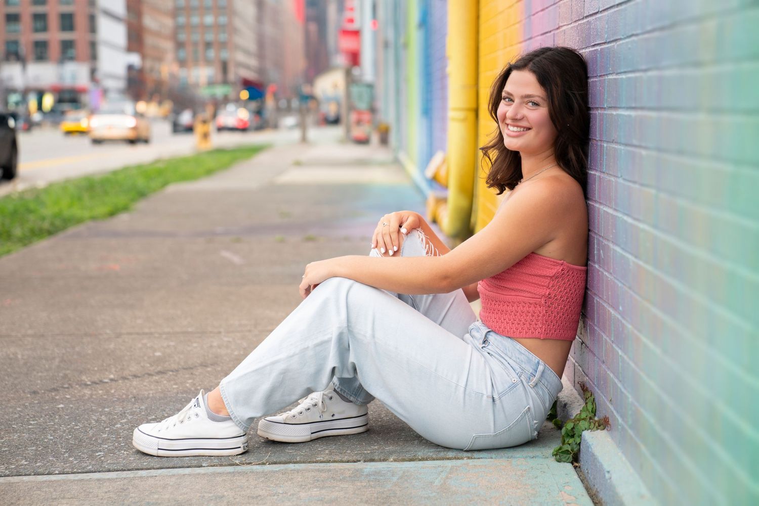 Person in white jeans sitting against a colorful wall while scrolling on their phone in an urban setting.