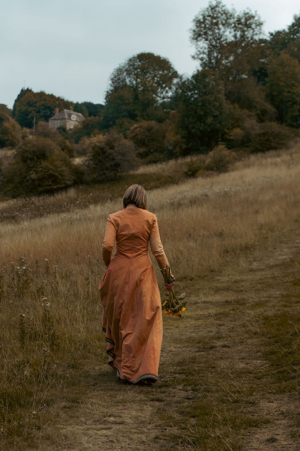 Author novel promotional photoshoot in Gloucestershire. A figure in a long orange dress walking on a hillside path while holding sunflowers.