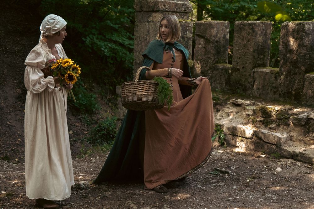 Author novel promotional photoshoot in Cotswolds. Two people in historical dress carrying baskets and flowers along a stone wall pathway.