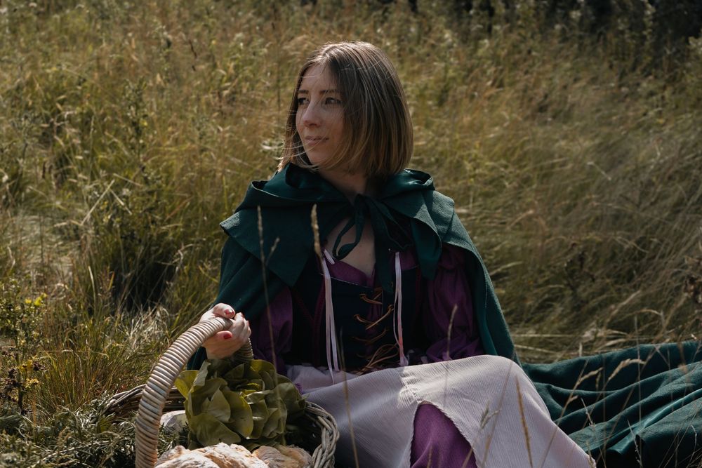 Author novel promotional photoshoot in Cotswolds. A person in medieval-style clothing sitting in a grassy field with autumn colors.