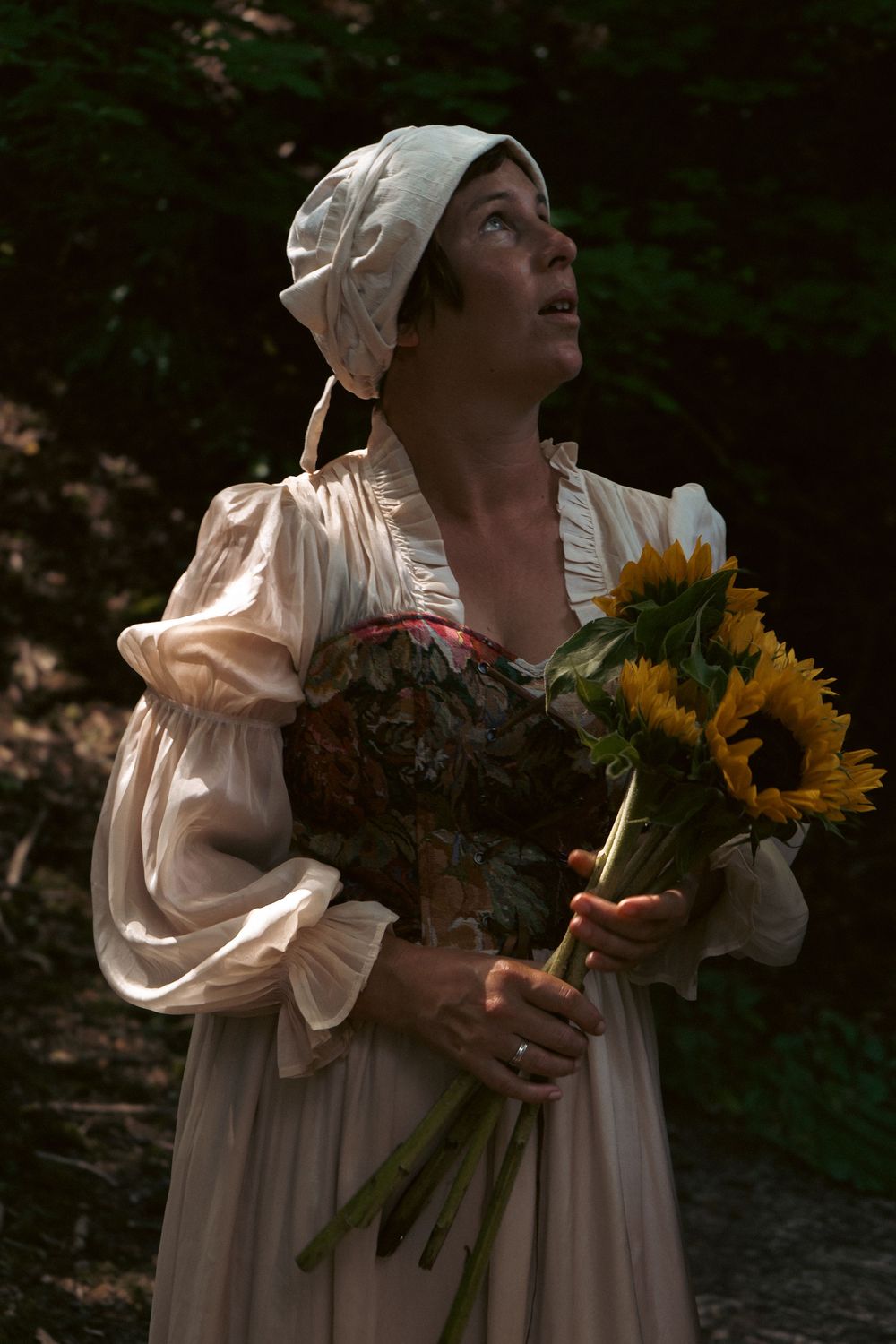 A person in historical costume holding bright yellow sunflowers in dramatic lighting. Author novel promotional photoshoot in Gloucestershire