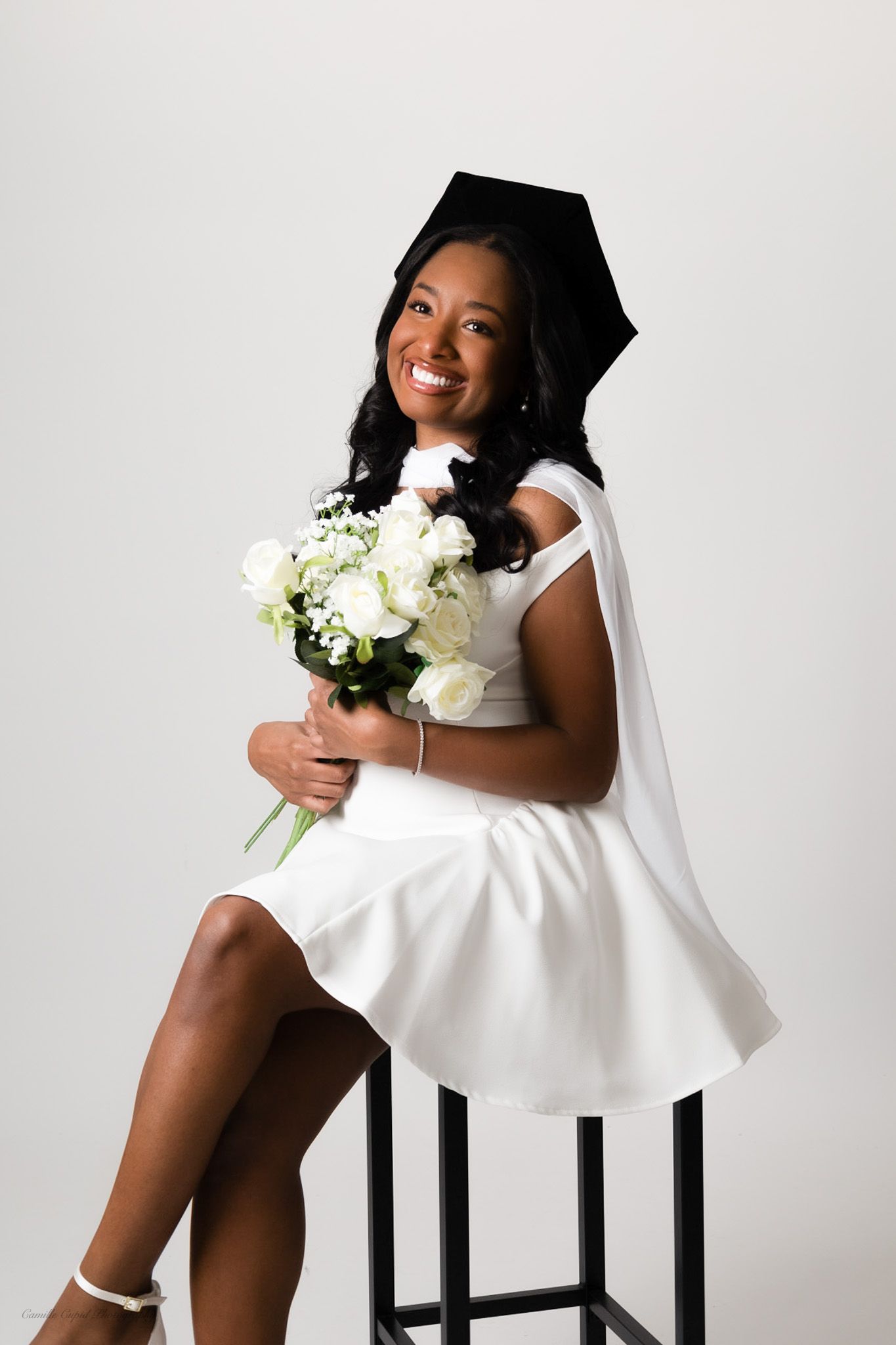 smiling in cap and gown white dress, holding bouquet for senior graduation photoshoot by Camille Cupid Photography in Pittsburgh